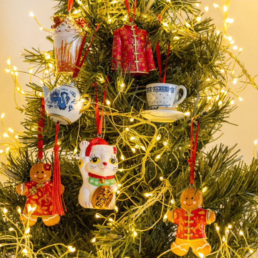 A set of colorful ceramic ornaments hanging on a Christmas tree with warm white string lights. The ornaments include a white cat with a santa hat and gingerbread men in red outfits, teacups, and a red jacket.