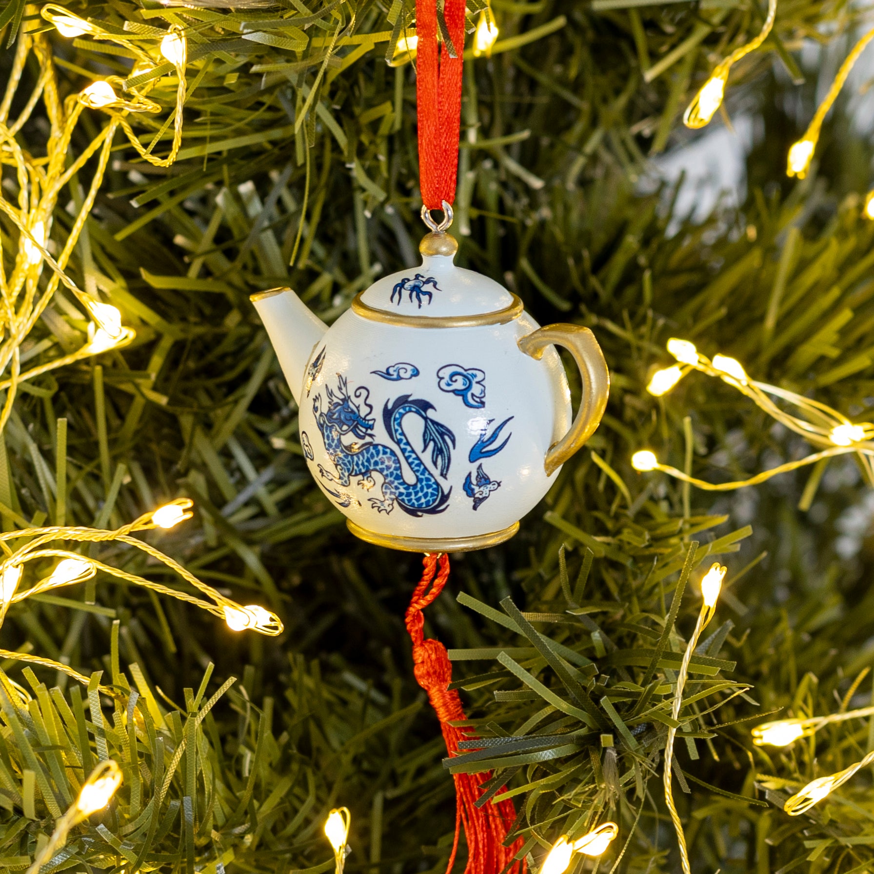A small white ceramic teapot ornament with blue dragon-style illustrations and gold trim, hanging from a red ribbon on a Christmas tree with warm string lights.