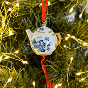 A small white ceramic teapot ornament with blue dragon-style illustrations and gold trim, hanging from a red ribbon on a Christmas tree with warm string lights.
