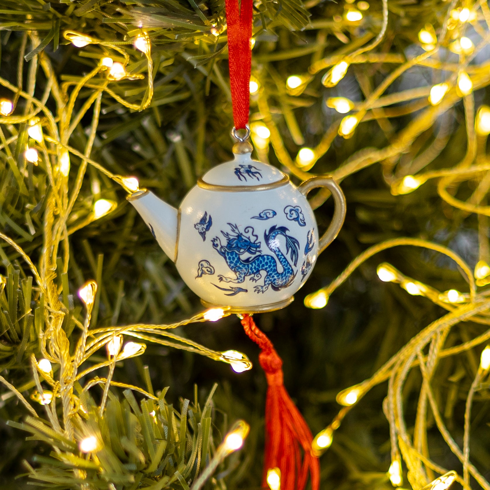 A small white ceramic teapot Christmas ornament with blue dragon illustrations and a red hanging ribbon, displayed on a green Christmas tree with warm yellow string lights.