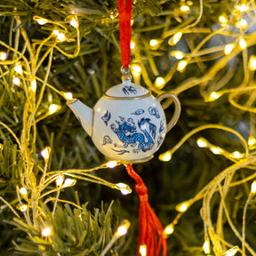 A small white ceramic teapot Christmas ornament with blue dragon illustrations and a red hanging ribbon, displayed on a green Christmas tree with warm yellow string lights.
