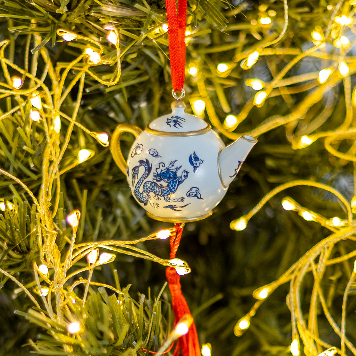 A small white ceramic teapot Christmas ornament with blue dragon and bird illustrations, hanging on a green Christmas tree with warm yellow string lights.
