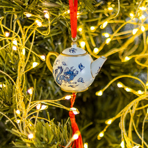 A small white ceramic teapot Christmas ornament with blue dragon and bird illustrations, hanging on a green Christmas tree with warm yellow string lights.