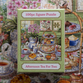 A 100-piece jigsaw puzzle box titled 'Afternoon Tea For Two' featuring an illustration of a tea-time scene with flowers, teapots, cups, and a three-tiered serving stand. The box is centered against a matching floral puzzle background.