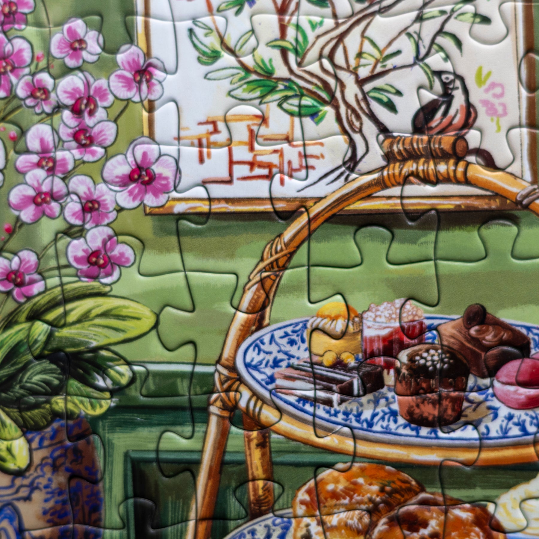 A close-up of a completed jigsaw puzzle showing a colorful still life scene with pink flowers, a green wall, a wooden chair, and assorted pastries on blue-and-white plates.