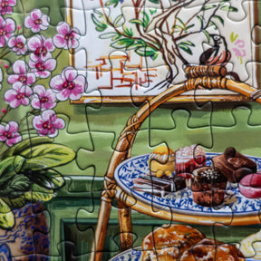 A close-up of a completed jigsaw puzzle showing a colorful still life scene with pink flowers, a green wall, a wooden chair, and assorted pastries on blue-and-white plates.
