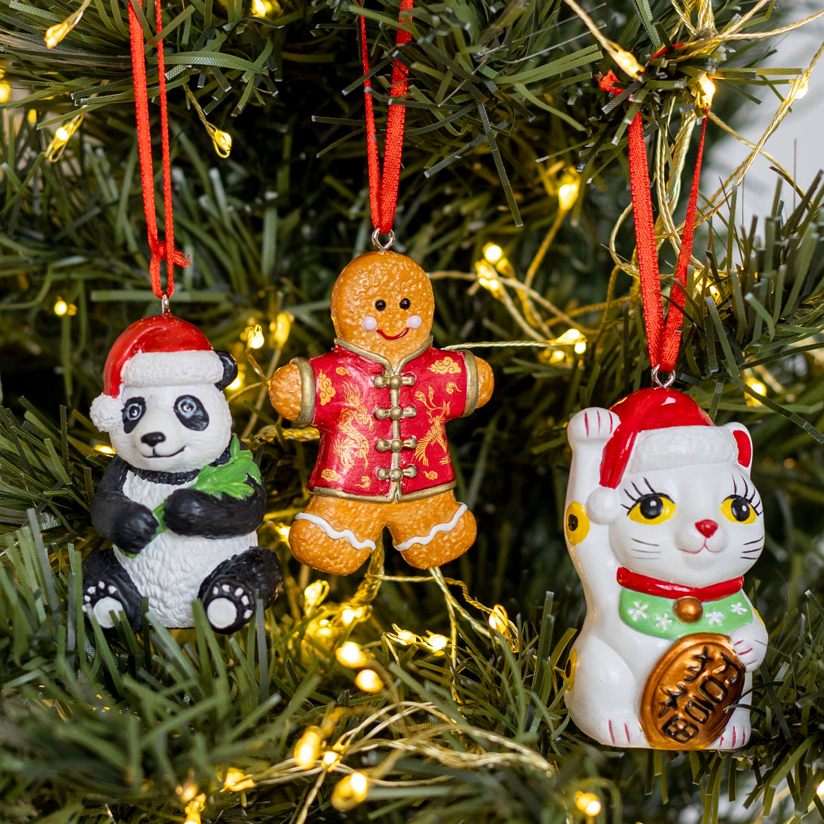 Three festive Christmas ornaments hanging on a lit Christmas tree: a panda wearing a Santa hat and green scarf, a gingerbread figure in a red Chinese-style outfit, and a white maneki-neko cat ornament with a red Santa hat and gold coin detail.