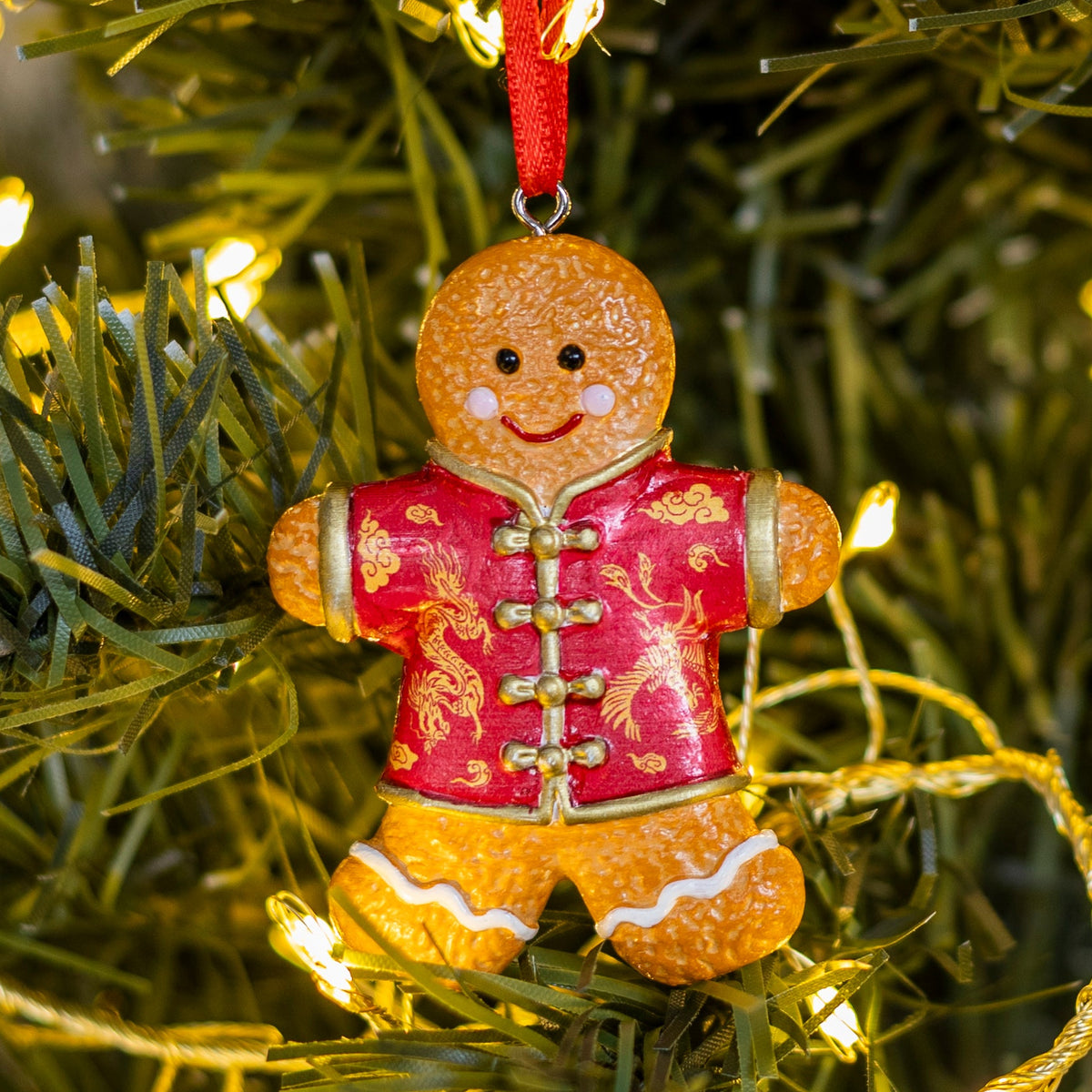 A gingerbread man Christmas ornament wearing a red Chinese-style jacket with gold dragon patterns, hanging on a green Christmas tree branch with warm string lights.