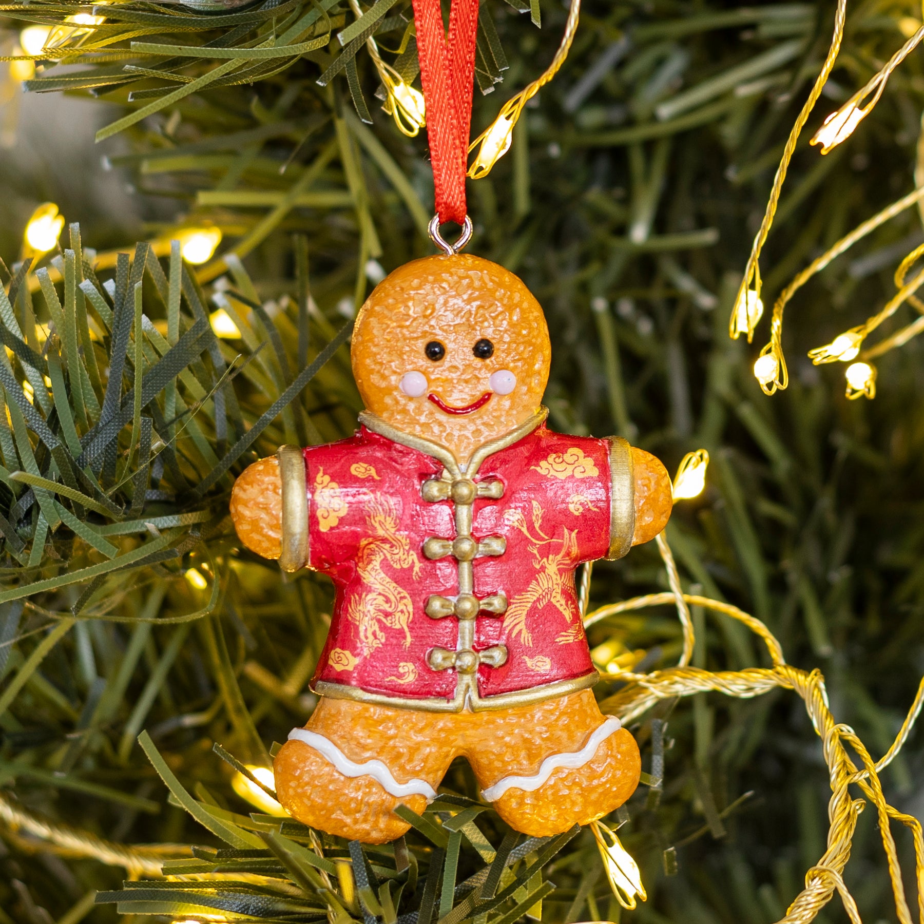 A gingerbread man Christmas ornament hanging on a green artificial tree with warm white string lights. The ornament is orange-brown with a red Chinese-style jacket featuring gold dragon motifs and gold buttons, and a red hanging ribbon.