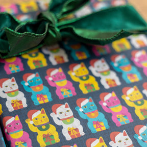 Close-up of wrapped gift boxes featuring wrapping paper with a repeating pattern of colorful maneki-neko cats on a dark background, tied with a dark green satin bow.