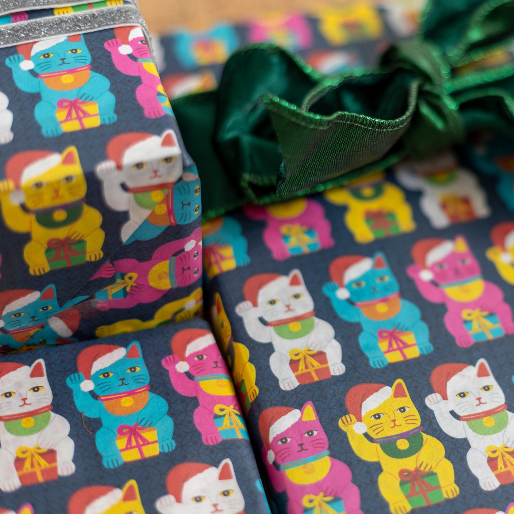 Close-up of wrapped gift boxes featuring wrapping paper with a repeating pattern of colorful lucky cats on a dark background, tied with a dark green satin bow.
